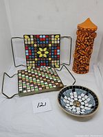 Front view of two square mosaic tile trivets with metal handles, small mosaic tile bowl, and tall orange carved flower candle