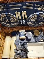 Photo showing the picnic basket interior with plates, mugs, thermoses, metal tray, flatware (forks and spoons), and cloth napkins.