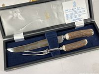 Carving knife and fork inside the lined storage box, showing knife blade and fork prongs with antler handles.