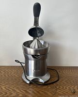 Front view of the Breville Citrus Press Pro showing the conical juicing cone, strainer, spout and the lever handle in upright position with power cord extending from base.