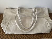 Front view of beige canvas tote bag with leather handles and bottom trim, laid flat on wooden surface.