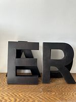 Photo of two large black metal letters 'E' and 'R' showing front sides and profile, placed on wooden surface against white wall.