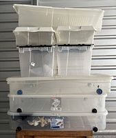 Stacked plastic storage containers of various sizes, predominantly white and transparent, some with wheels, with contents visible in the lowest transparent bin