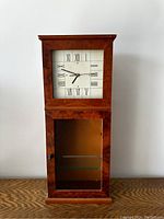 Front view of the rectangular vintage wall clock with Roman numeral face and lower glass door with knob revealing a glass shelf inside.