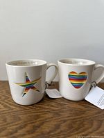 Two white ceramic coffee mugs with rainbow-colored star and heart cut-outs displayed side by side on a wooden surface.