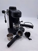 Front and side view of Krups Mini espresso machine with glass frothing pitcher, portafilter, filter basket, and frothing nozzle placed on white surface.