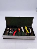 Opened metal tackle box showing multiple fishing lures in green felt compartments.