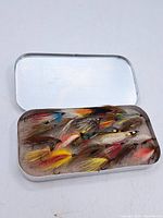 Closed metal case open to show assortment of vintage fishing flies with multiple colors and feather types