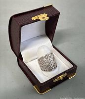 Sterling silver filigree ring shown on white padded display inside brown textured ring box with gold clasp, angled view.