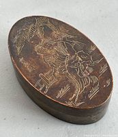Oval metal trinket box with engraved Chinese figures and landscape on the lid, angled top view showing engraving details and tarnish.