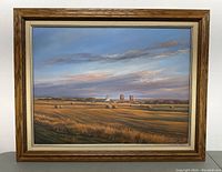 Framed landscape painting with fields, hay bales, and silos under a sky with clouds and sunset tones.