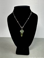 Full view of the necklace displayed on a black bust, showing the silver chain with spaced beads and two pendants with green stones.