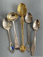 Five sterling silver spoons laid out, largest with ornate gold-tone and engraved bowl, medium sized ladle, and three smaller decorative spoons including an enameled spoon.