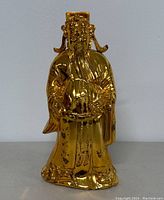 Front view of gold-tone metal figurine coin bank showing a traditional robed figure holding a fan with a few discolored spots on surface.