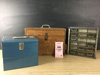 Three industrial storage boxes arranged side by side on a wooden surface against dark background: from left to right, blue metal box, wooden toolbox, and metal/ plastic parts sorter.