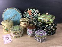 Full view of 9 vintage tins arranged together showing variety in shapes, sizes, and designs, including floral and scenic motifs.