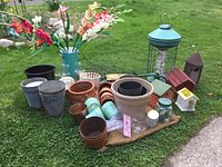 Overview of assorted garden items on grass including large green bird feeder, many plant pots of various sizes and types, decorative bird houses, artificial gladiolus flowers in vase