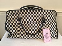 Front view of black and white checkered weekend bag placed against a white wall, showing black vinyl handles, front pocket and zipper top closure.