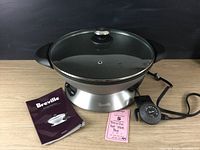 Full frontal view of the Breville Hot Wok Pro on its electric base with glass lid, temperature controller and instruction book laid out on wooden surface.