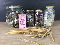 Four glass jars containing assorted buttons placed behind a group of bamboo knitting needles, showcasing the variety of buttons and needles together.
