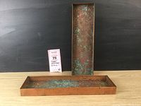 Pair of rectangular copper plant trays arranged with one standing vertically and the other lying horizontally on a wood surface with a dark background. Both trays show green verdigris patina on the surface.