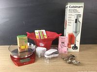 Full lot showing all items: Cuisinart hand blender in box, red kitchen scale, popcorn popper, measuring spoons, chip clips, can opener, and sealed box of Food Huggers.