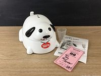 Front view of Hero Hot Dog Steamer shaped like a white dog with black ears and nose, showing smiling face and tongue, with measuring cup and instruction manual beside it