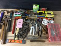 Full assortment of hand tools and hardware items arranged on table including hammers, screwdrivers, pliers, measuring tapes, level, rasp file and red organizer box with small metal hardware.