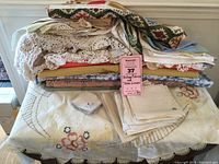 Folded assorted fabrics in various prints and textures stacked with embroidered linens underneath; includes large floral embroidered tablecloth with crocheted edge visible