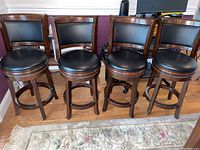 Four wooden bar stools with padded black vinyl seats and backs, showing the full set in a row.