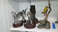 Six bird figurines displayed on a shelf, showcasing pewter material and a colorful goldfinch with naturalistic base