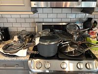 Overall view of cookware lot stacked on stove and counter, including pots, pans, and lids