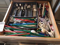 Full drawer view showing assorted cutlery including forks, spoons, knives, and colorful plastic-handled utensils and straws/stirrers