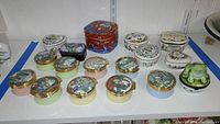 Overview photo of the collection on a white shelf showing mostly round enameled pill boxes with scenes, some heart shaped and novelty dishes.