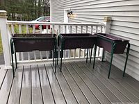 Three raised rectangular plant/flower beds on green metal stands displayed on a porch deck; two are empty, one contains soil and dried plants.