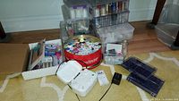 Wide view of sewing supplies on floor featuring a white bobbin winder, round tin with threads, various plastic boxes with threads and bobbins, power adapter, and small containers with sewing accessories.