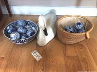 Photo of white ceramic Montana Decoys swan figurine, blue and white decorative ceramic balls in bowl and wicker basket on wooden floor with scale reference sticker.