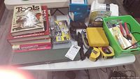 Photo of the lot on table showing books, tools, measuring tapes, sheathed knives and red handled pliers