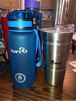 Blue Ion BPA Free water bottle with black and blue cap and blue carrying strap next to stainless steel Bubba tumbler with clear lid on a wood table.