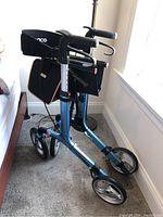 Folded view of the blue-frame portable wheelchair with visible ergonomic handles and frame structure, placed indoors on carpet.