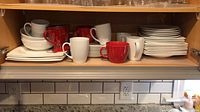 Shelf with stacked white plates and bowls along with white and red mugs behind