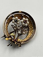 Front view showing hammered circle frame, seed pearl flowers with ruby and emerald centers, ribbon bow accent