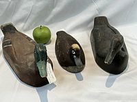 Three wooden duck decoys displayed on white cloth with a green apple for size reference. The decoys show wear and have painted details. One is larger with greenish-blue head, others smaller with dark brown and cream colors.