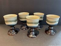 Five pottery goblets arranged on a dark surface with a dark background, showing their front view with colored bands and glossy stems.
