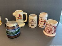 Five beer steins and mugs arranged side by side on a flat surface, showing diverse designs and markings.