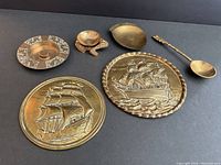Photo showing entire lot of 6 vintage brass items arranged on a black background. Includes 2 ship-themed round plaques, decorative spoon, frog figure, coin dish, and brass base.