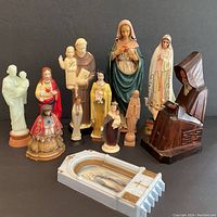 Photo showing all 12 religious figurines arranged on a black background, includes wooden monk figure, various Virgin Mary and saint figures mostly resin.