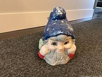 Front view of old concrete garden gnome showing facial details, blue hat, white beard, and red jacket with signs of paint wear.