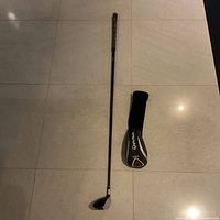 Full view of the golf club and TaylorMade branded black head cover on tile floor.