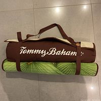 Rolled up mat with attached brown pillow, showing Tommy Bahama logo on pillow and green palm leaf print on mat.